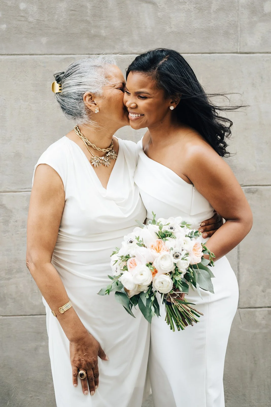 Two women, one elder and one younger, wearing white dresses, embrace in front of a concrete wall. The younger woman holds a bouquet of white and peach flowers with green foliage. The elder woman has gray hair in a bun with a yellow hair clip and is wearing jewelry. The younger woman has long black hair and is smiling.