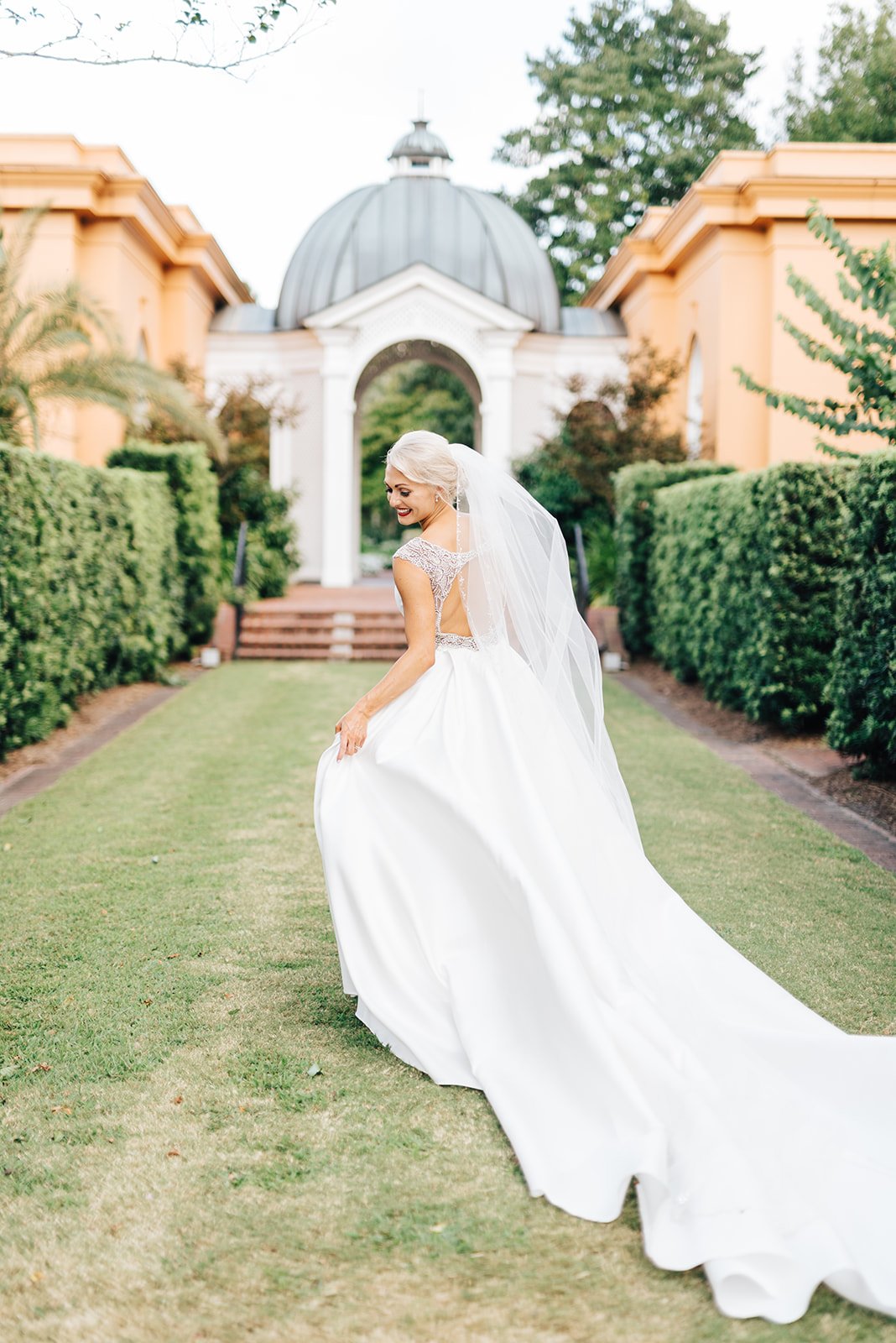 A bride in a white wedding dress with a long train, smiling and looking over her shoulder, standing on a grassy path surrounded by greenery, with a building that has an arched entrance and a dome in the background.