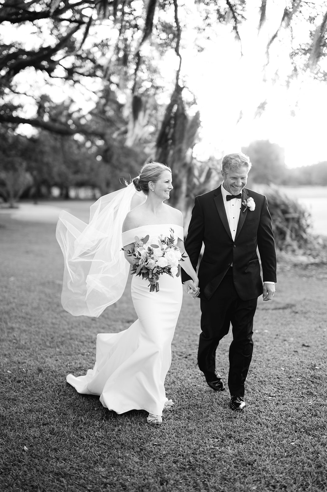 A bride and groom walking hand-in-hand outdoors, smiling, with trees in the background, captured in black and white.