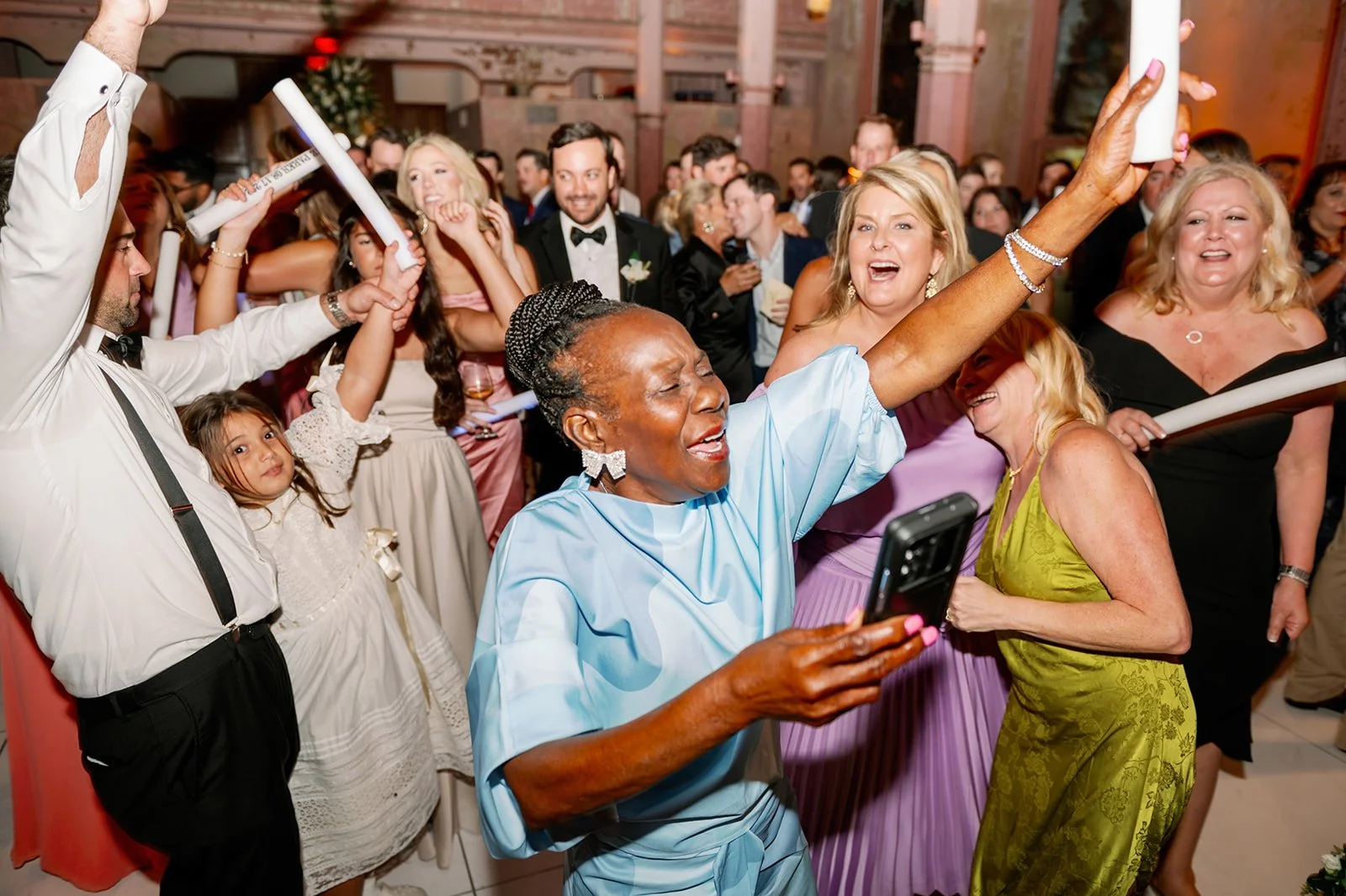 A lively celebration with diverse people dancing, smiling, and taking selfies at a wedding or party in a decorated venue.