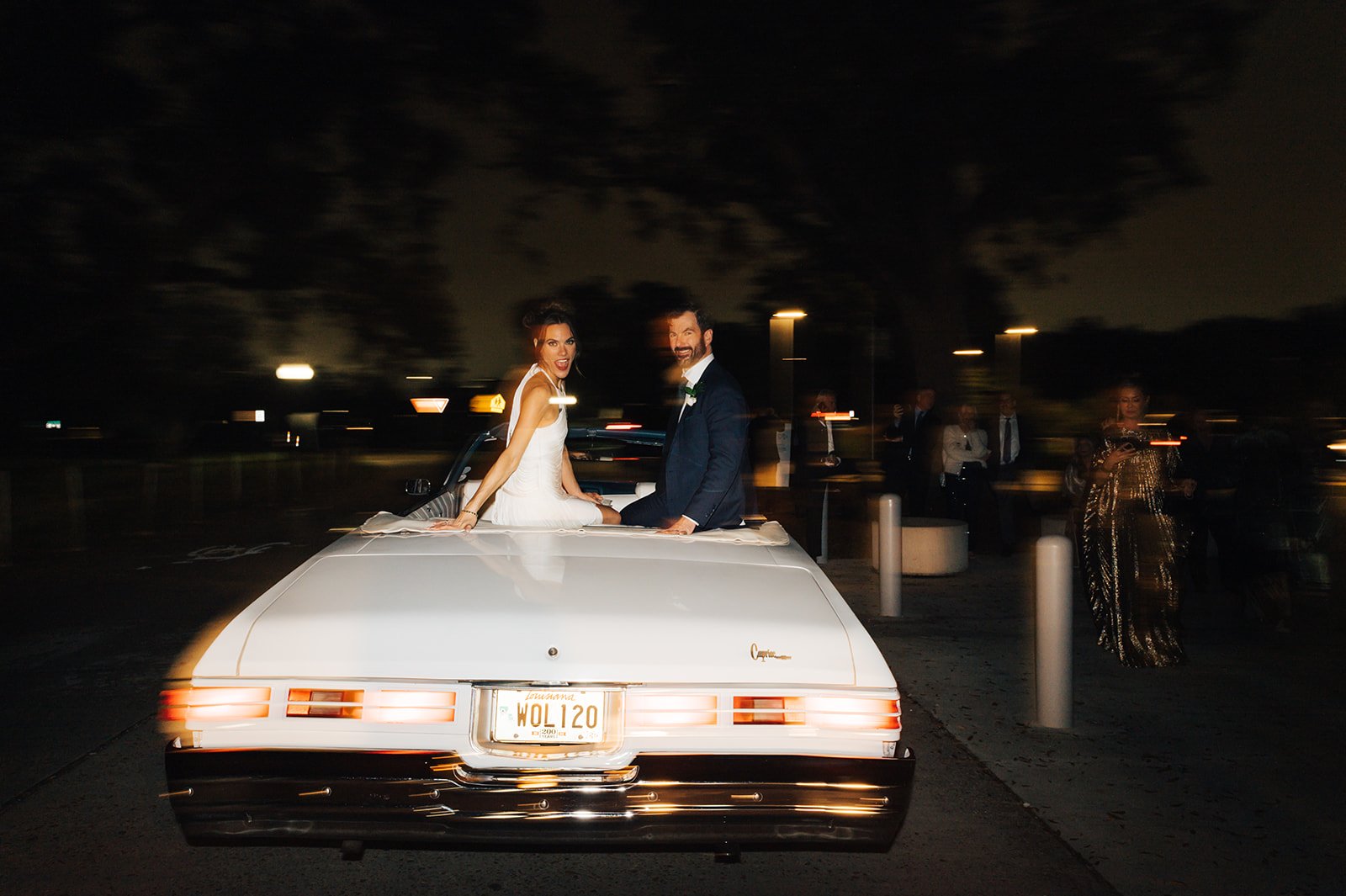 Newlywed couple sitting on the back of a white limousine during their wedding celebration at night, with wedding guests in the background.
