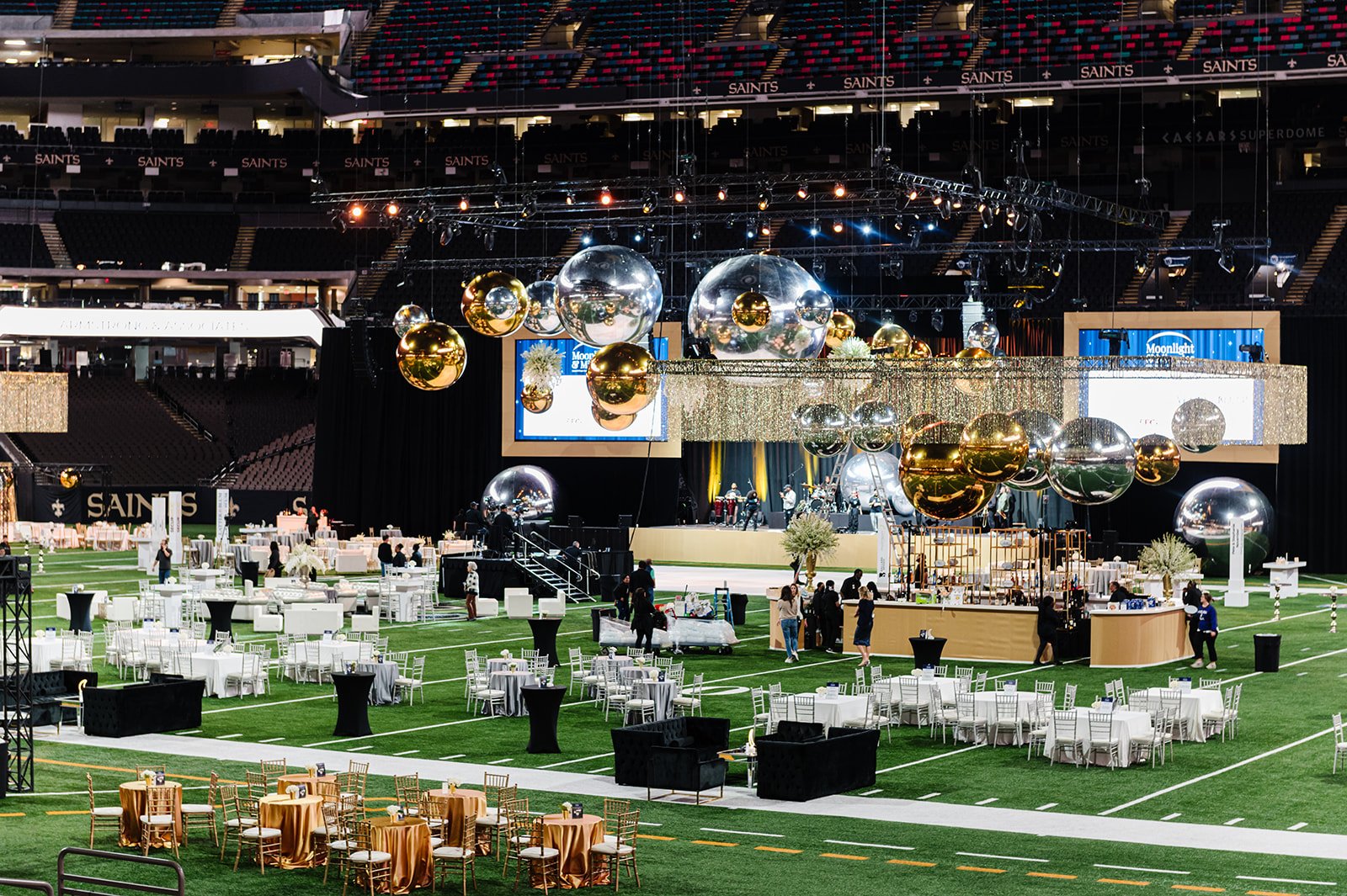 Event setup inside a stadium with decorated tables, balloons, a stage with a band, and large screens, preparing for a celebration.