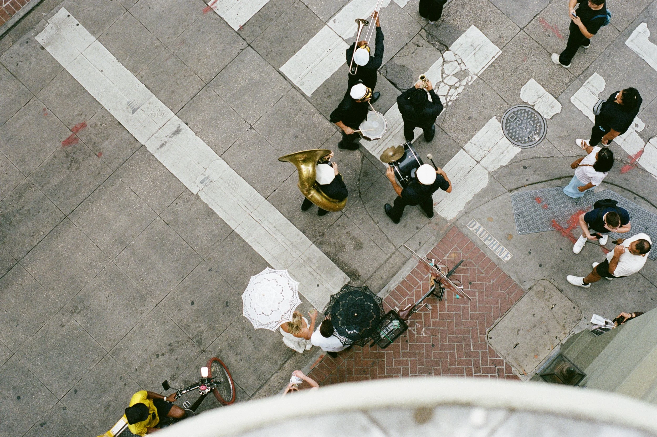 A bird's eye view of a city crosswalk showing a musical band playing, several pedestrians, and umbrellas on a sidewalk.