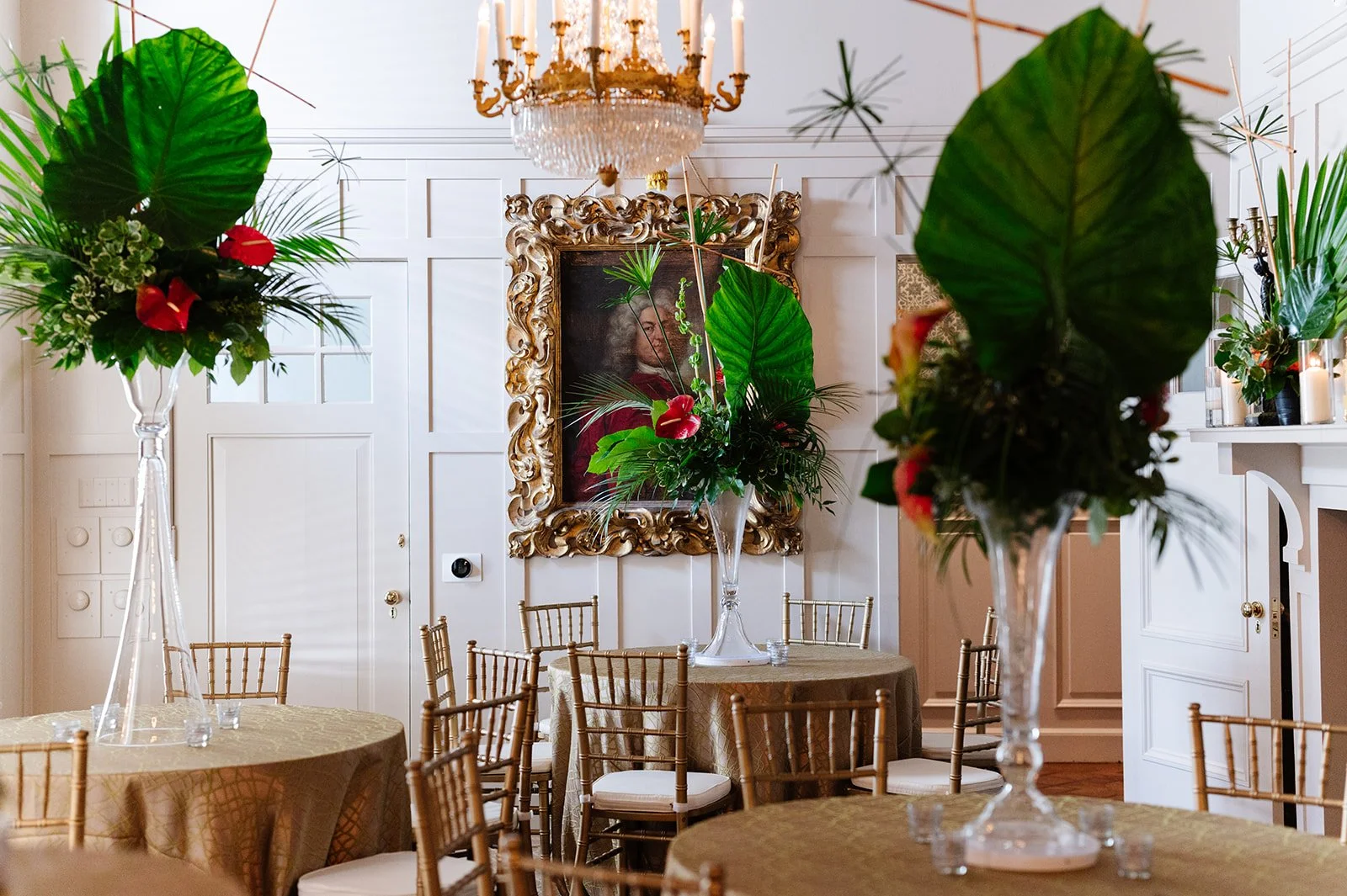 Elegant event space with tables covered in gold tablecloths, tall glass vases with tropical floral arrangements, a large ornate framed portrait on the wall, and a chandelier hanging from the ceiling.