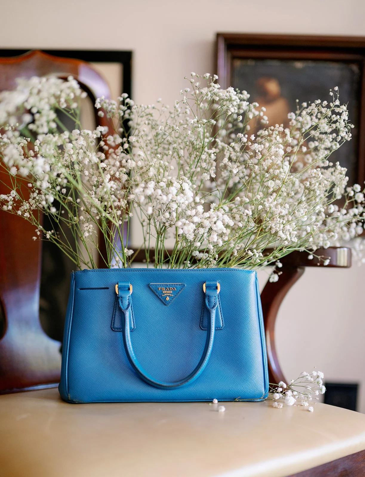 A blue Prada handbag filled with white flowers, placed on a wooden surface with a wooden mirror frame and a painting in the background.