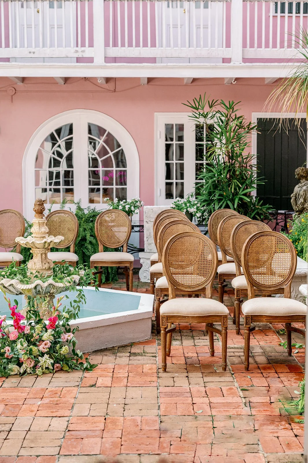 A garden patio with a pink wall, white arched window, surrounded by green plants, wooden dining chairs with beige cushions, a small white fountain, and a black door, set on a brick floor.