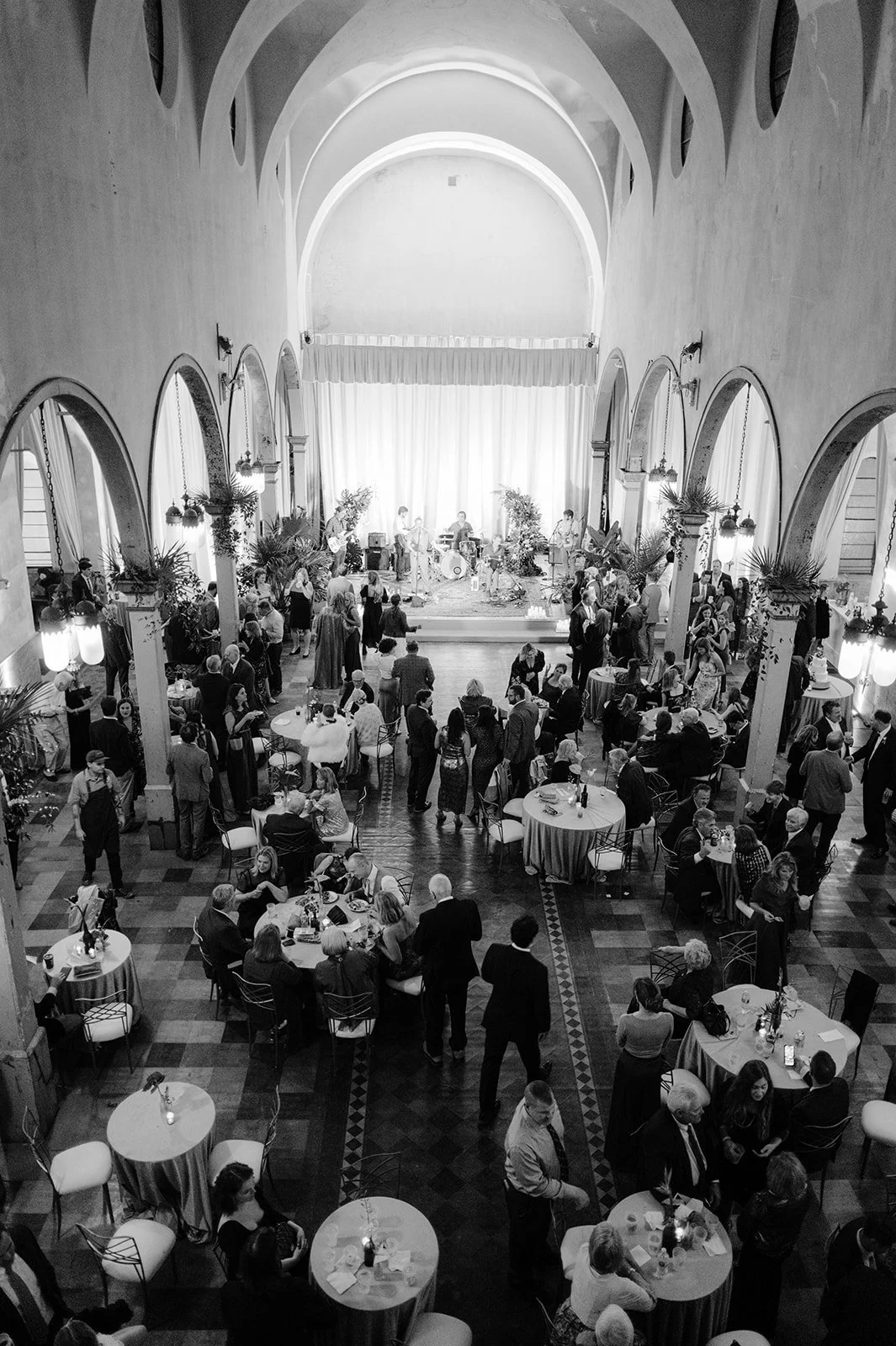 Black and white photo of a lively indoor celebration with many people socializing and dancing, a band playing on stage at the front of the room, and tables set with candles and decorations.
