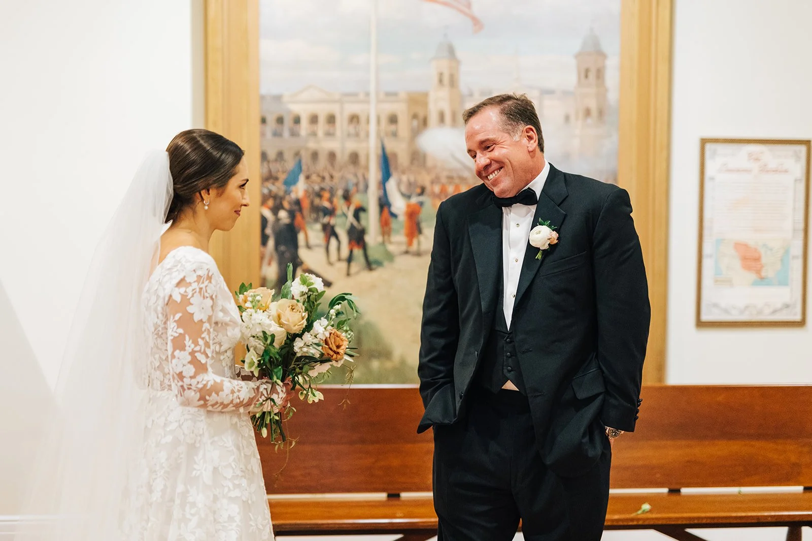 A bride in a lace wedding dress and veil holding a bouquet of flowers, smiling and looking at a groom in a tuxedo with a boutonniere, indoors with a backdrop of a historic painting and a map.