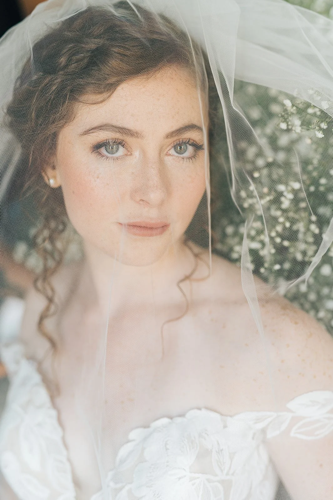 Close-up of a young bride with curled hair, wearing a lace wedding dress and veil, looking softly at the camera.