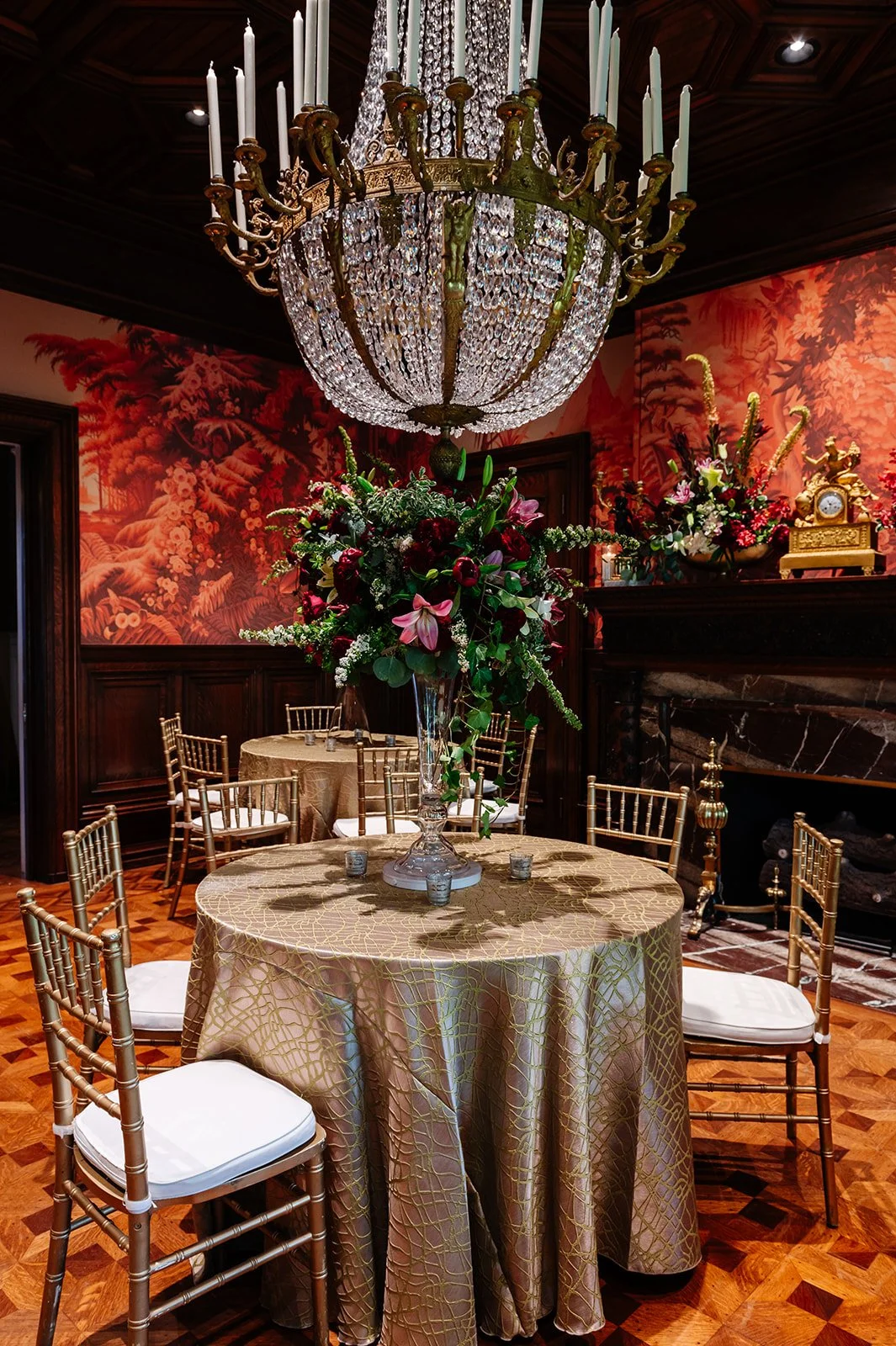 A luxurious dining room with a chandelier, a large floral centerpiece on a round table, surrounded by gold chairs, with a red scenic wallpaper and a fireplace in the background.