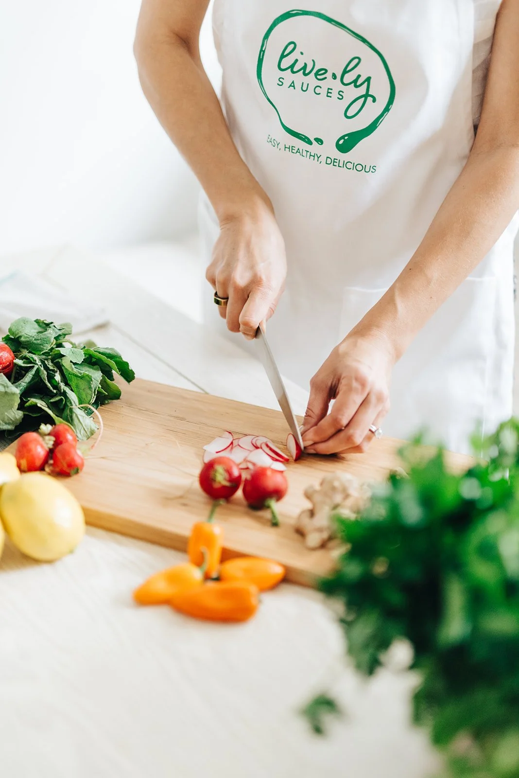 Person slicing radishes on a wooden cutting board surrounded by fresh vegetables like strawberries, lemons, ginger, and peppers.