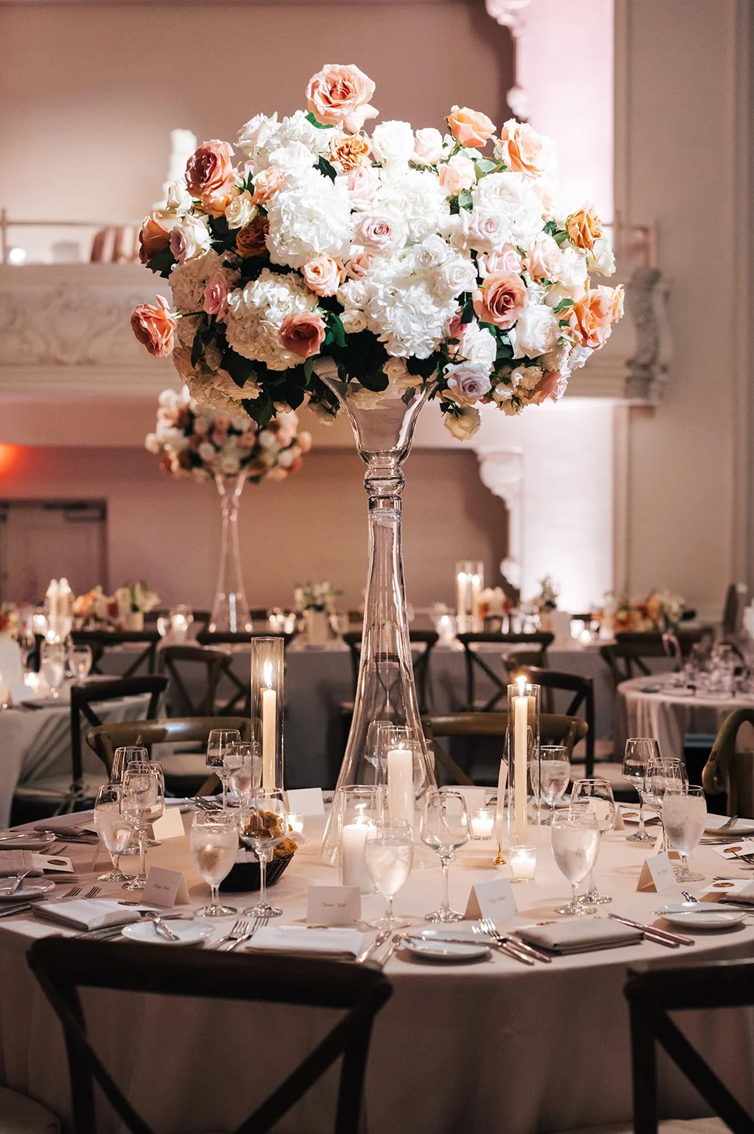 Elegant wedding reception table with a tall glass vase holding a large bouquet of white and peach roses and hydrangeas, surrounded by lit candles and set with glassware, cutlery, and place cards.