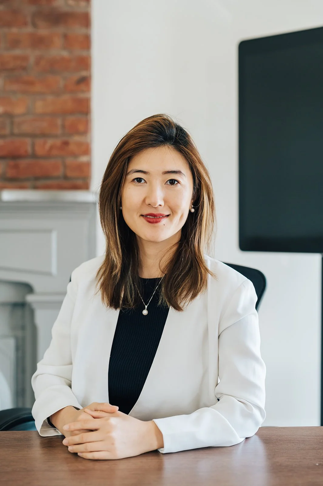 A woman with shoulder-length brown hair, wearing a white blazer and black top, sitting at a wooden table in an office setting with a brick wall and a large black monitor behind her.