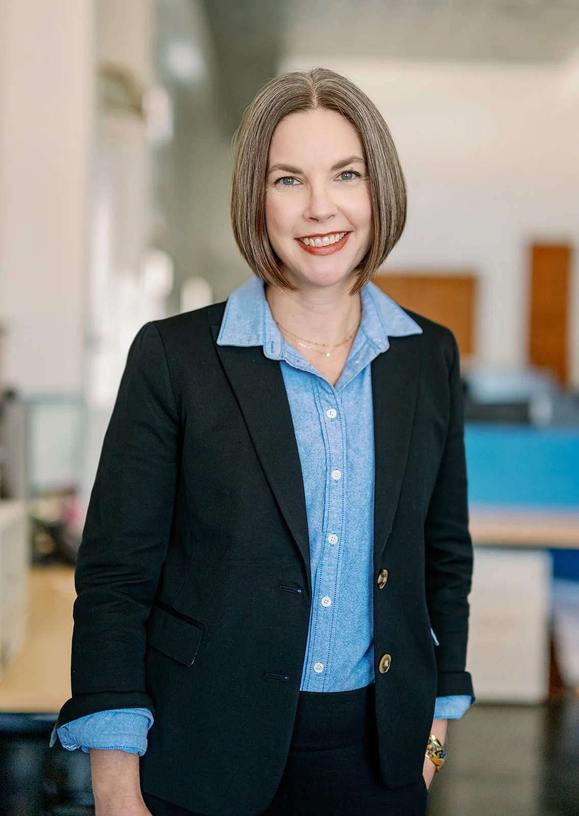 A woman with short brown hair, smiling, wearing a black blazer over a blue button-up shirt, standing indoors with a blurred background.