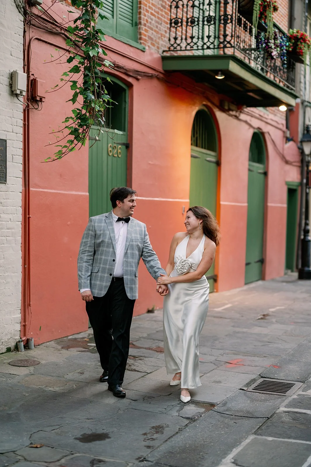 A couple walking hand-in-hand on a city street, smiling and looking at each other, with colorful buildings and a balcony in the background.