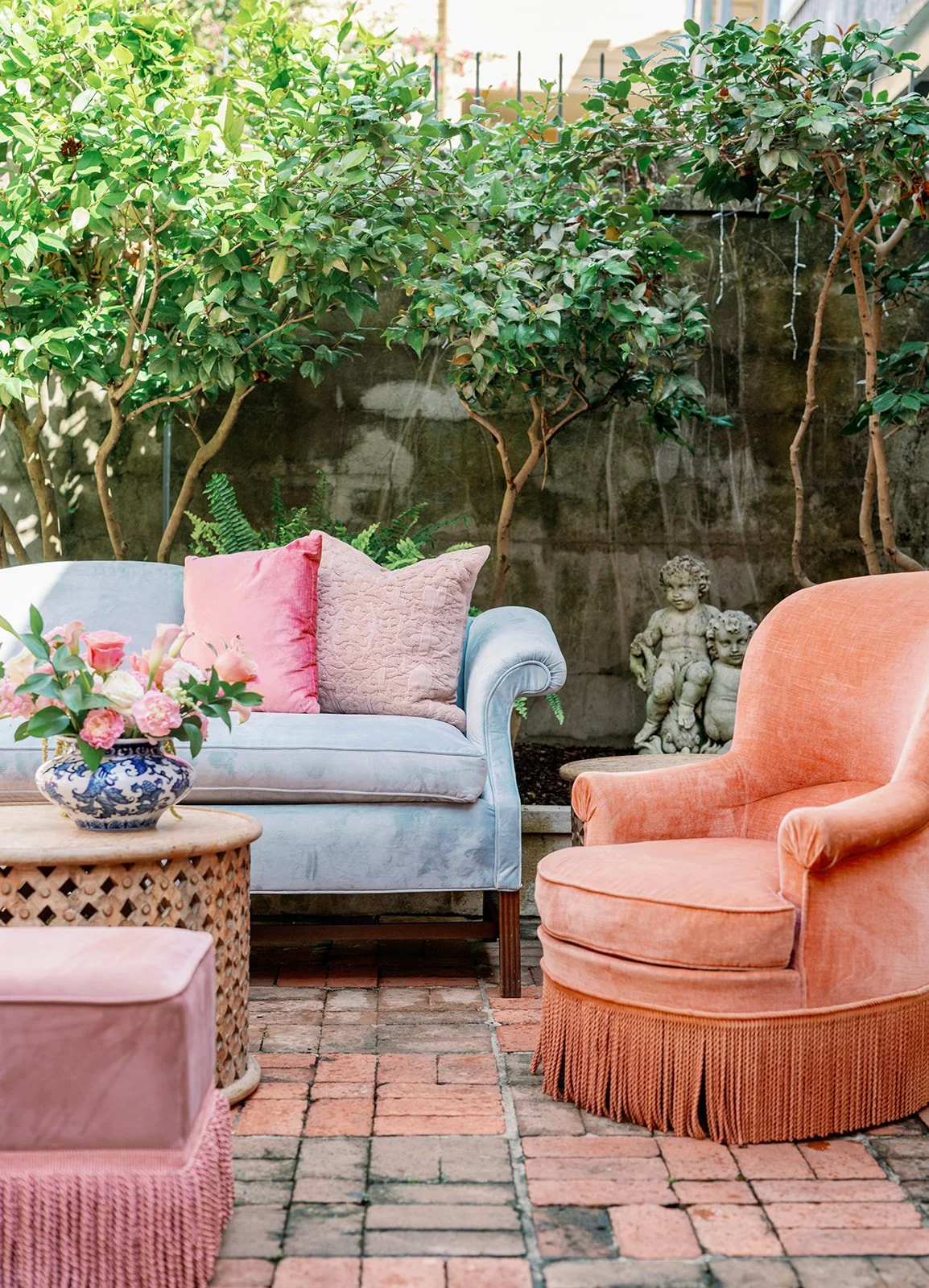 A cozy outdoor patio with a light blue sofa adorned with pink and textured pillows, a peach armchair, a rattan coffee table with pink flowers, and lush green trees in the background.