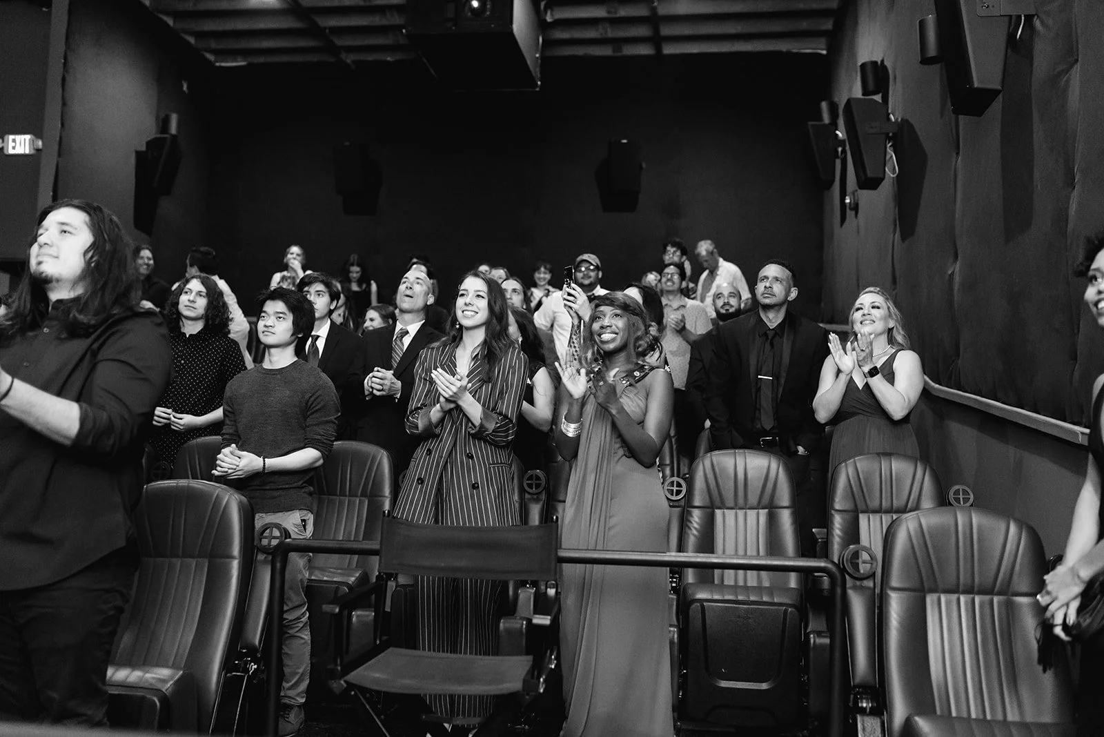 Audience standing and clapping in a dimly lit theater or concert hall.