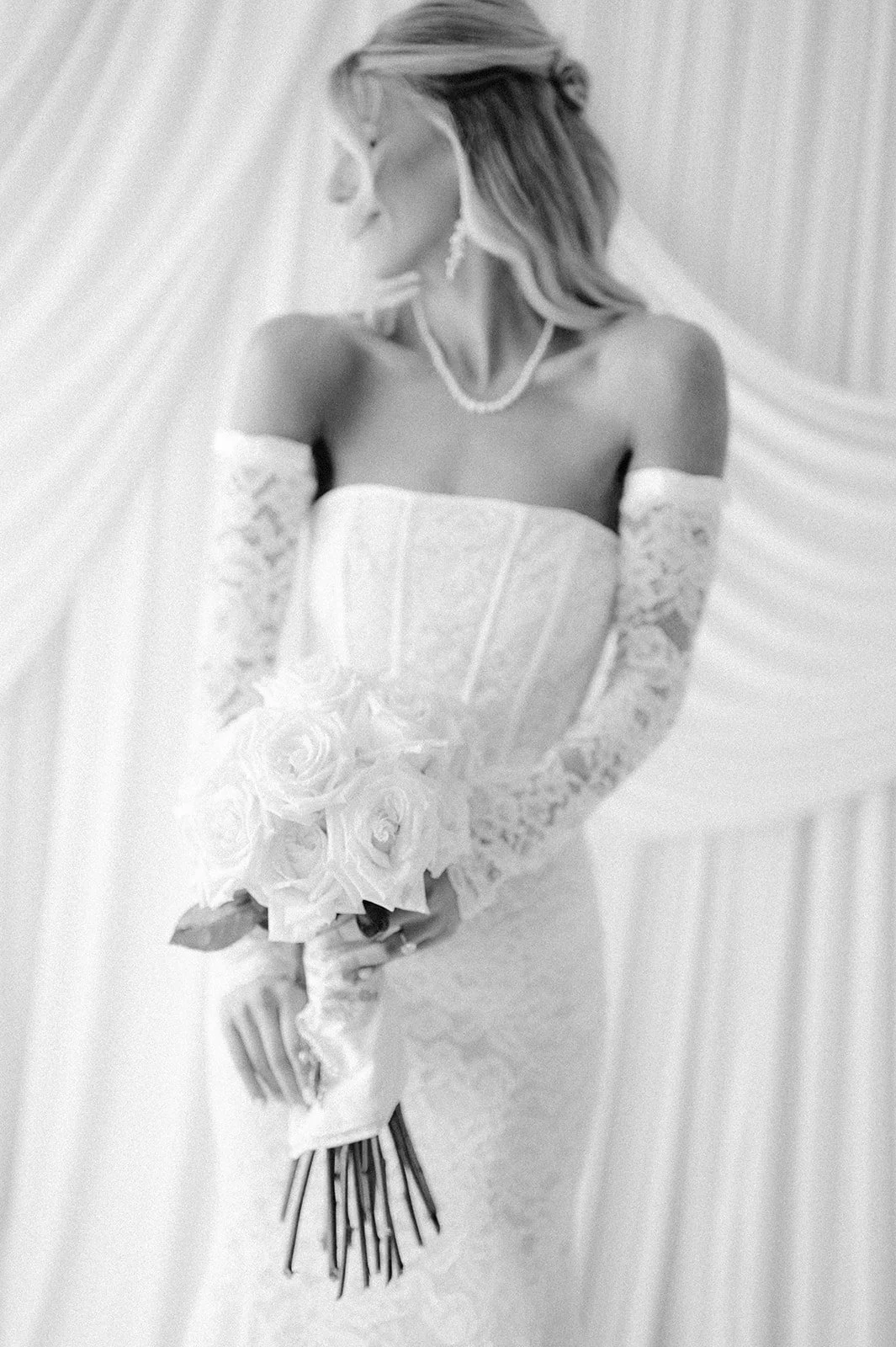 A woman in a wedding gown holding a bouquet of roses, dressed in lace gloves and jewelry, standing against a draped curtain background.