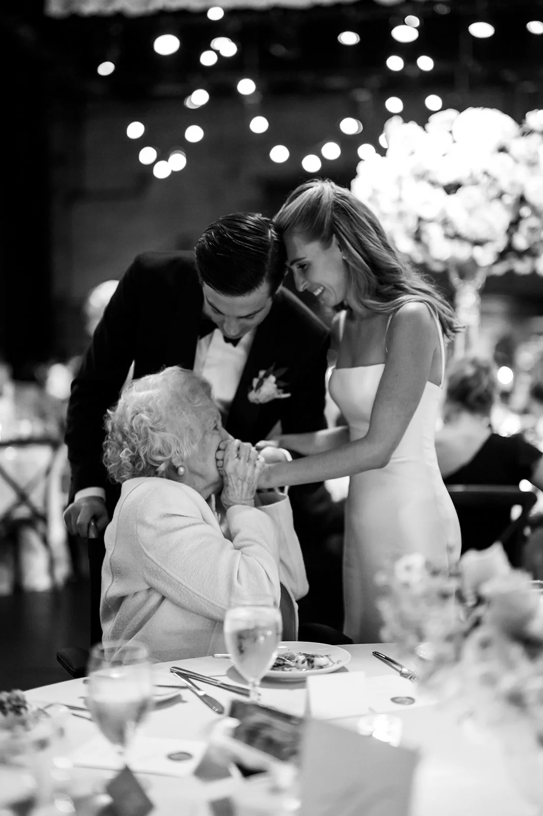 A wedding reception scene in black and white, showing a bride and groom leaning over an elderly woman, smiling and holding hands, with festive lights and floral decorations in the background.