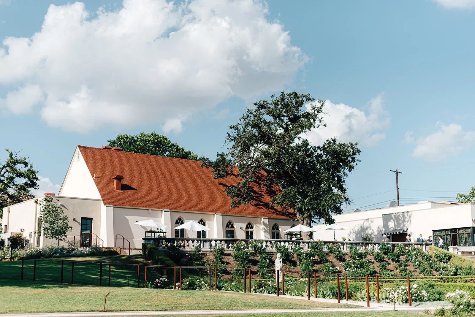A white building with a red-tiled roof, surrounded by trees, a lawn, and a garden with shrubs and flowers. There are people outside near umbrellas on a patio.