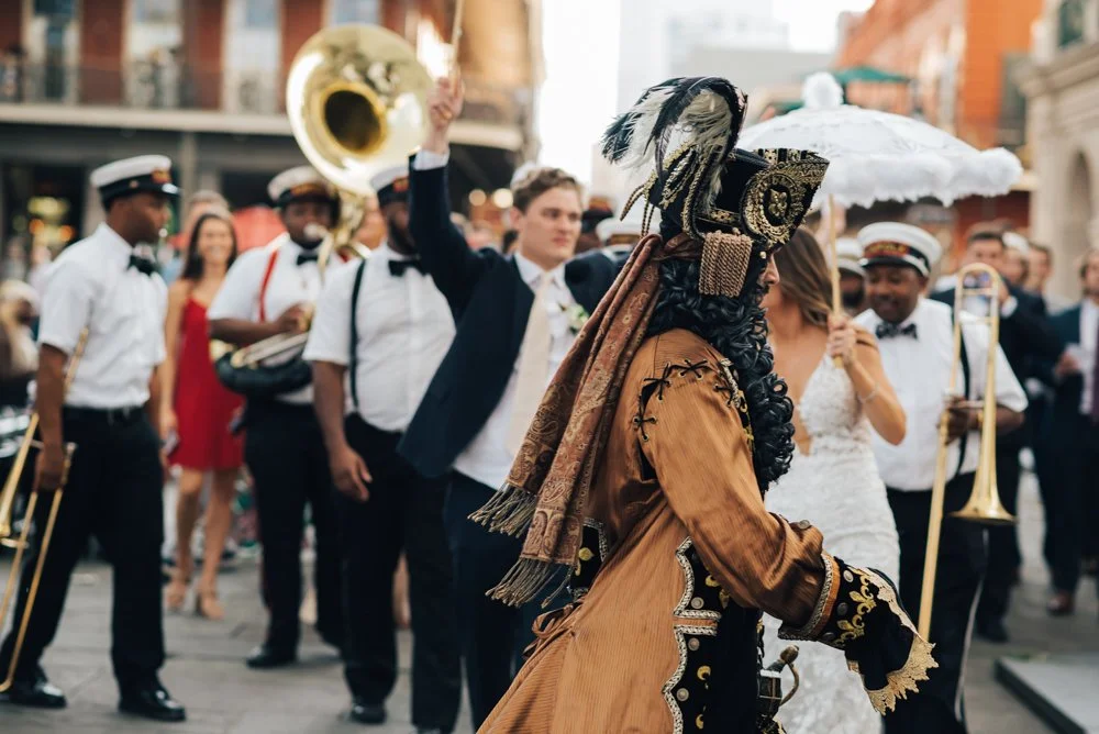 A parade with people in costumes, including a man in a vintage-style outfit with an elaborate hat, and a band playing instruments.
