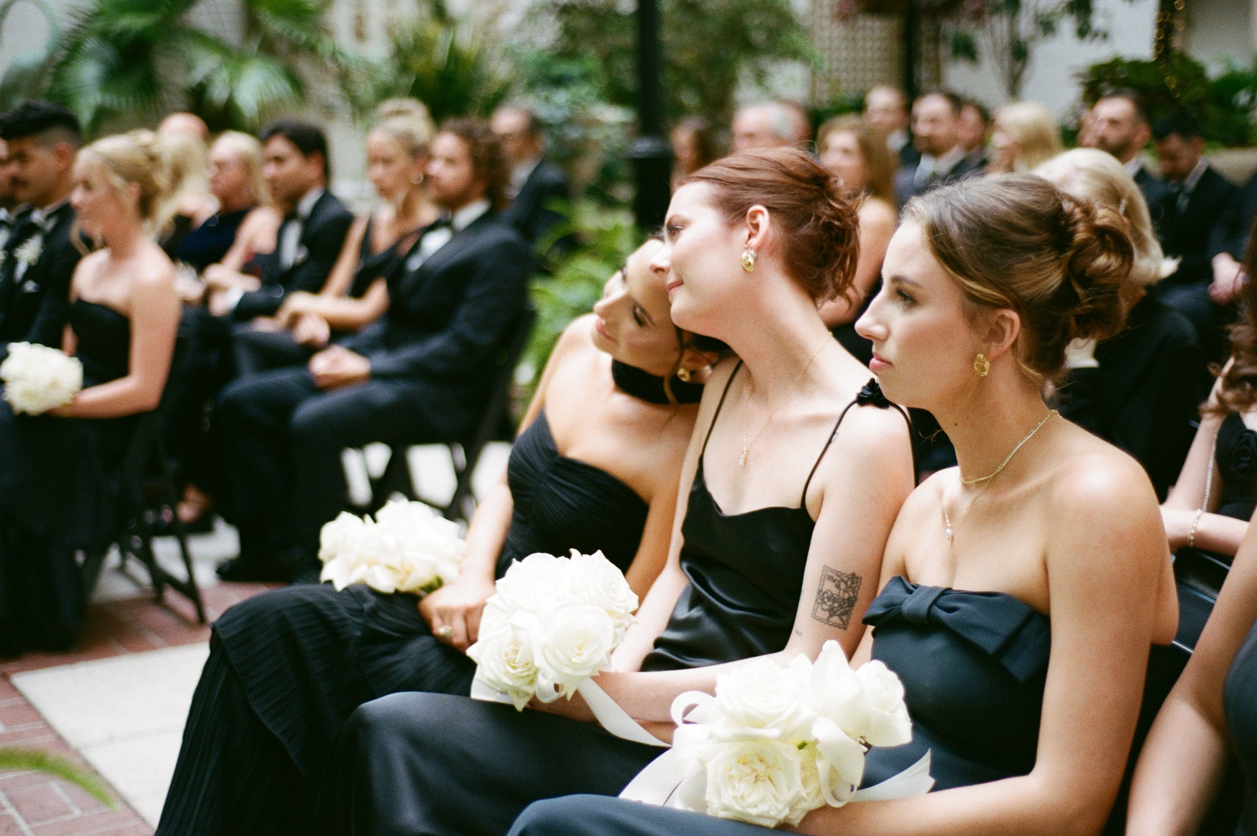 Group of women in black dresses, holding white bouquets, attending a wedding ceremony indoors with greenery in the background.
