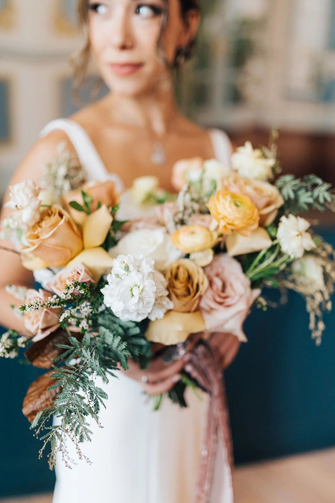 A woman in a white dress holding a large bouquet of mixed flowers including roses, ranunculus, and greenery, with a blurred indoor background.