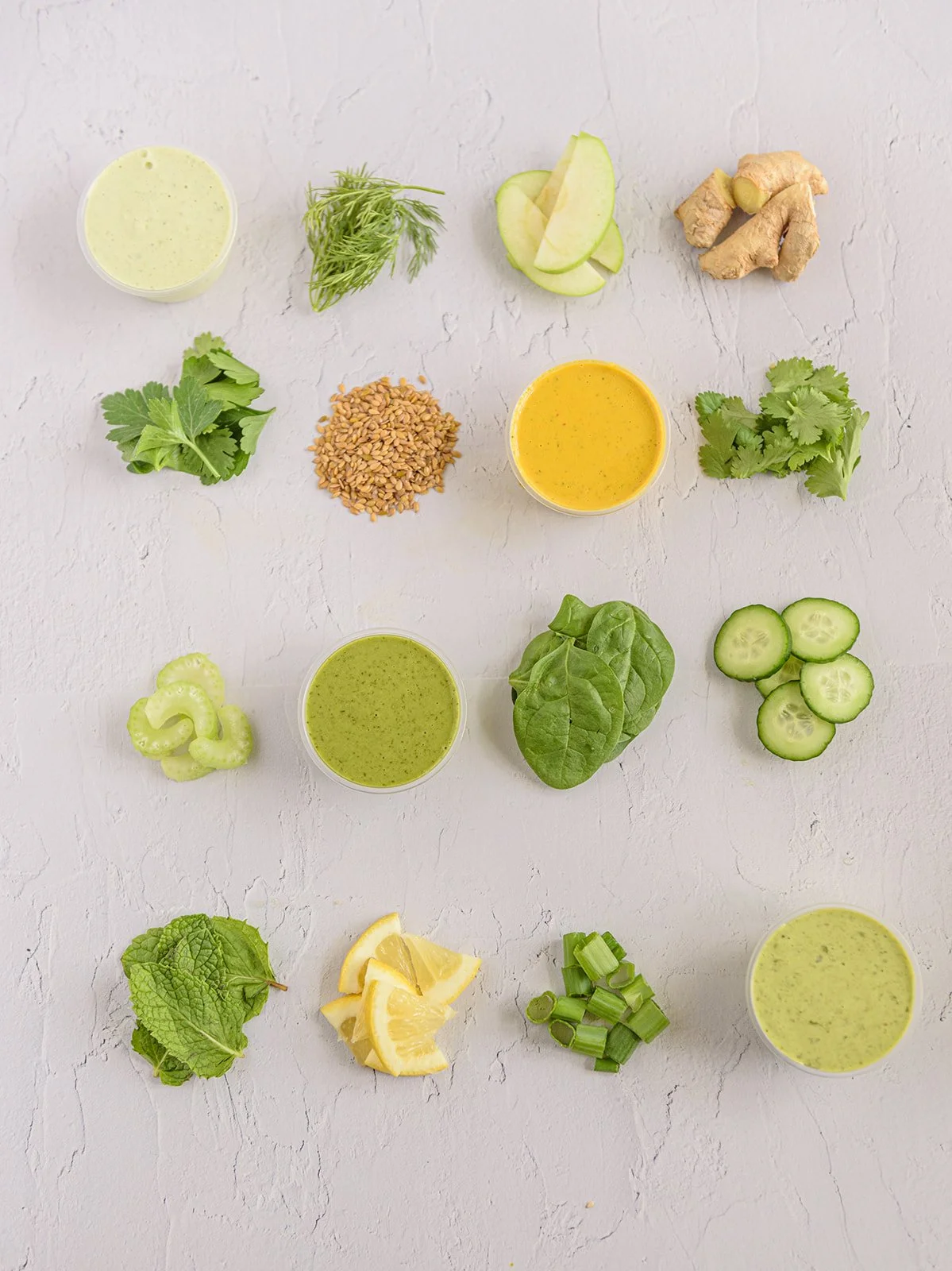 Arrangement of ingredients including ginger, diced apple, cilantro, sunflower seeds, cucumber slices, spinach, lemon wedges, green onions, and four different green sauces on a white textured surface.
