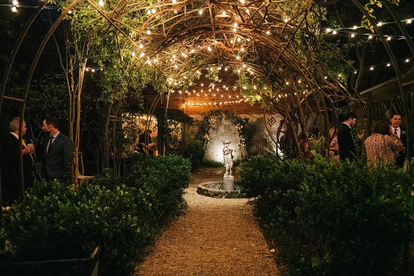 Nighttime social gathering under a lit garden arbor with string lights, a pathway leading to a fountain, and people in formal attire mingling in a lush outdoor setting.
