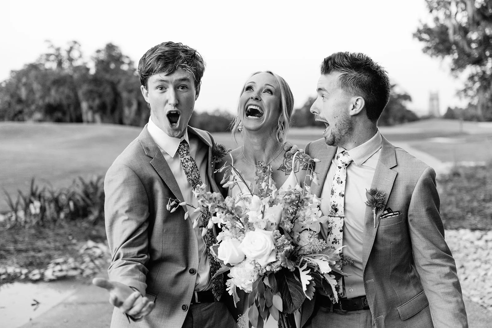 Three people, two men and one woman, dressed in suits and a dress, are outside, appearing excited or surprised, with a woman holding a bouquet of flowers, in a park or garden setting.