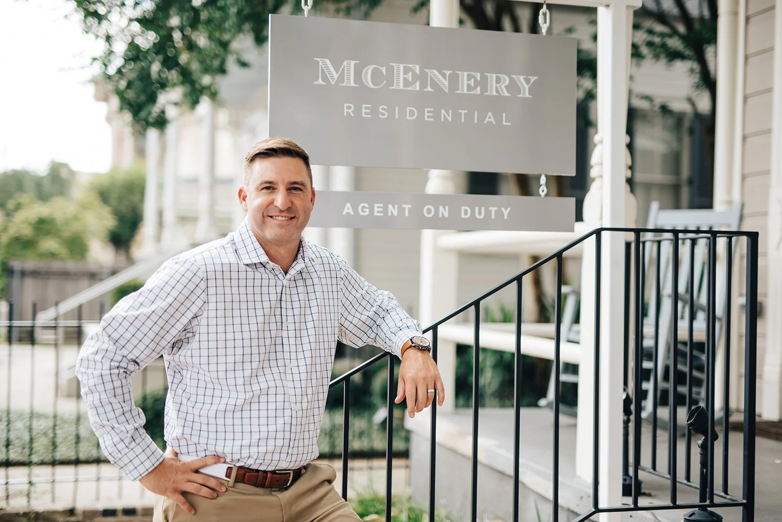 A man standing on the porch of a residential building, smiling and leaning on a black railing, with a sign behind him reading 'McEnery Residential' and 'Agent on Duty'.