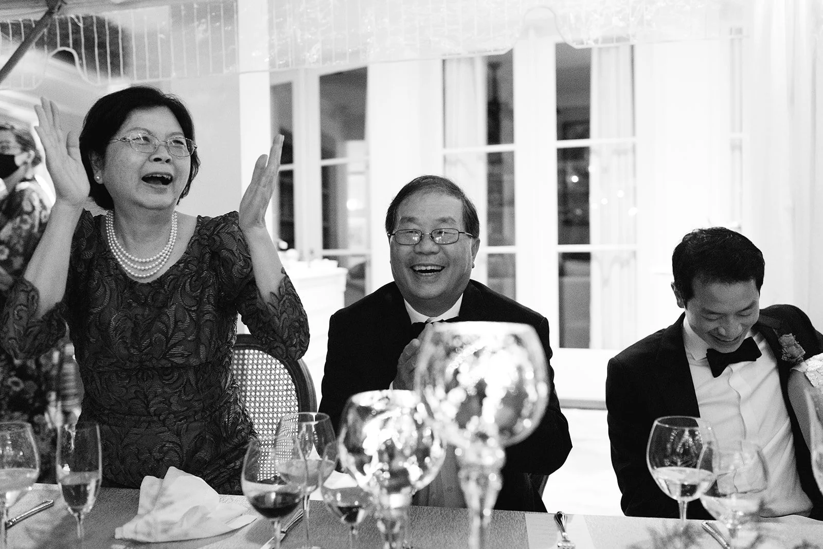Black and white photo of three people at a dinner party, smiling and laughing. A woman on the left is wearing glasses, a pearl necklace, and a lace dress. The man in the middle, wearing glasses and a tuxedo, has his hands clasped and is smiling. The 