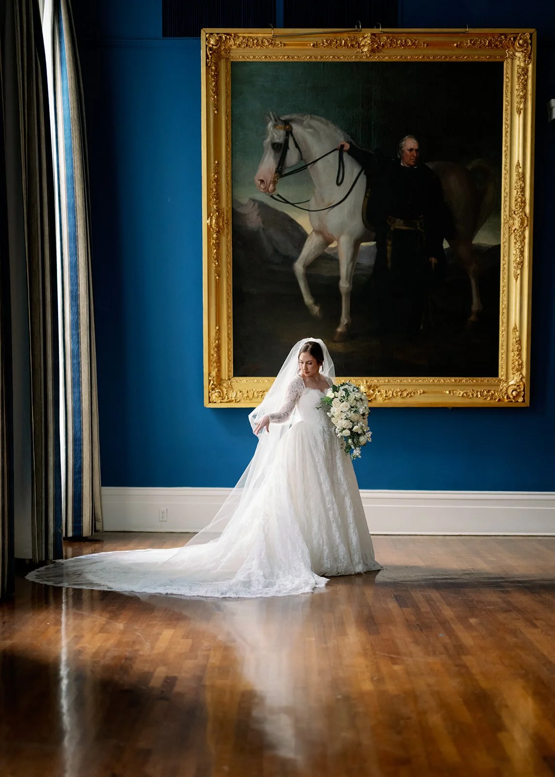 A bride in a white wedding dress and veil holding a bouquet of white flowers standing in a room with hardwood floors, large framed picture of a man on a white horse in the background, and blue walls.