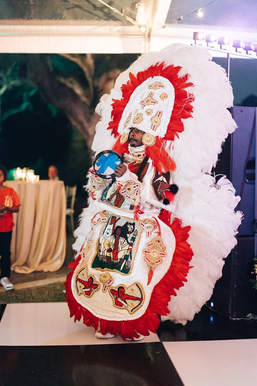 A performer in a colorful, elaborate costume with feathered headdress and embroidered cloth performs at an event. The costume features red and white feathers, and detailed artwork depicting cultural scenes, and the performer is holding a musical inst