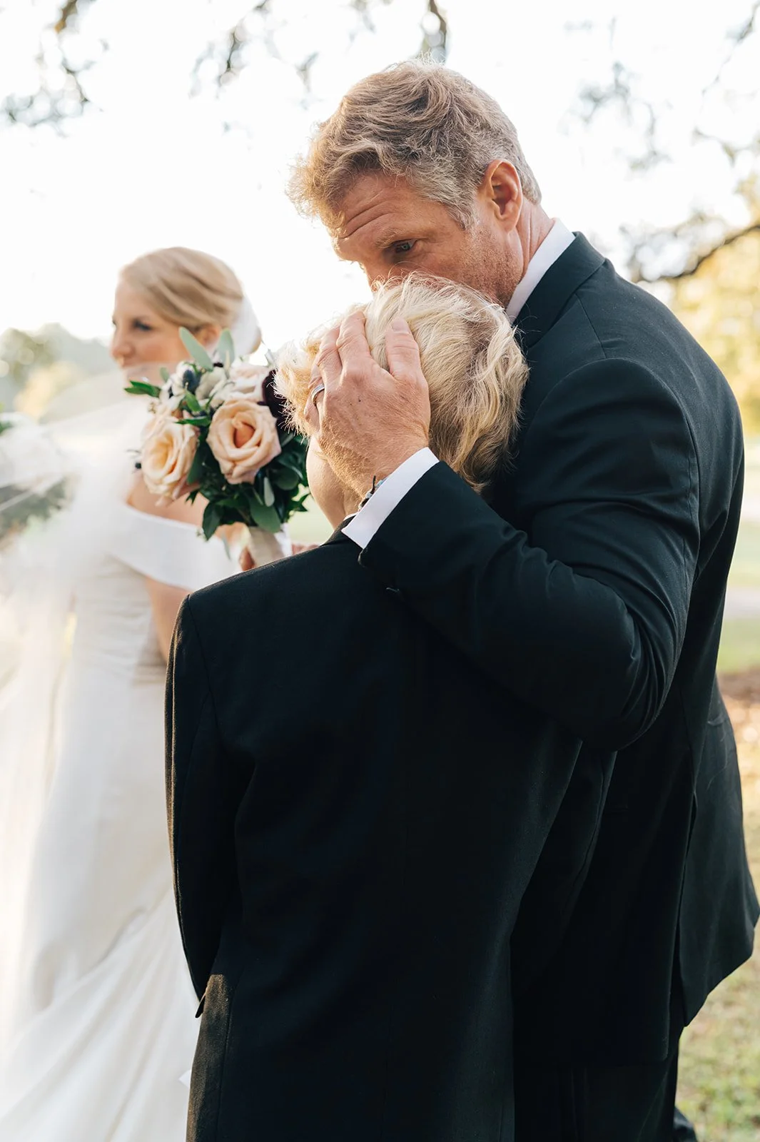 A young man dressed in a suit embraces an elderly woman, likely his mother, during a wedding ceremony outdoors. A bride holding a bouquet of flowers looks on in the background.