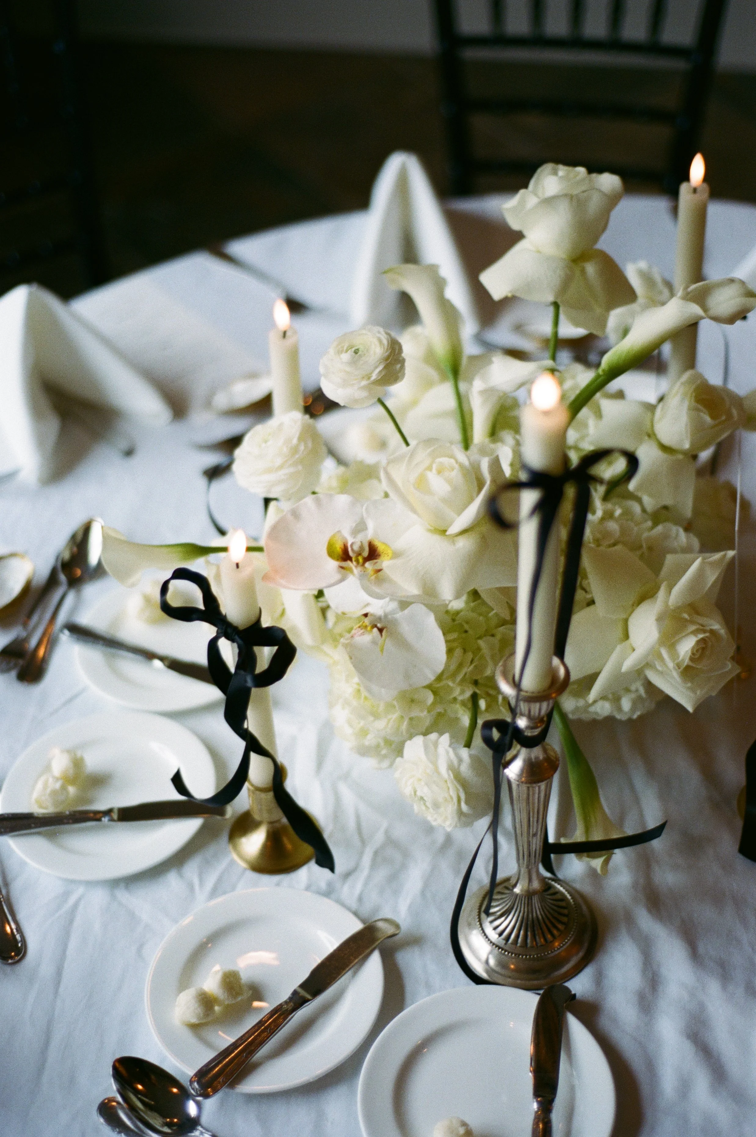 A table decorated with white flowers, candles with black ribbons, white tablecloth, and place settings with silverware and small plates.