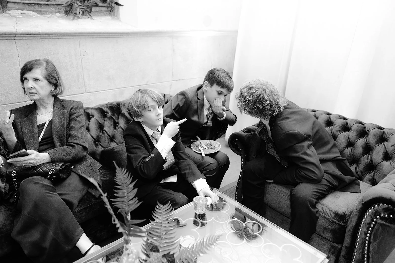 Four people sitting on a cushioned sofa having a conversation. Two boys in suits are in the middle, one is pointing and the other covers his mouth. Two women are on either side, one with a handbag and phone, the other leaning forward.