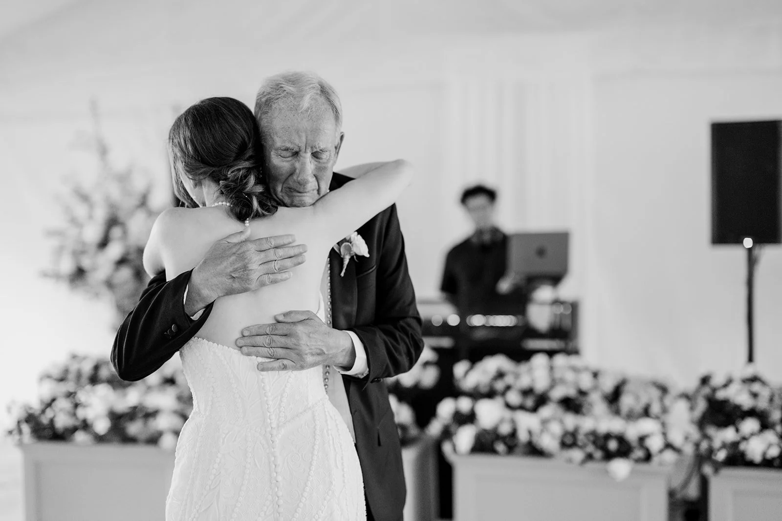 A bride hugging an elderly man, possibly her father, during a wedding reception with a musician playing in the background.