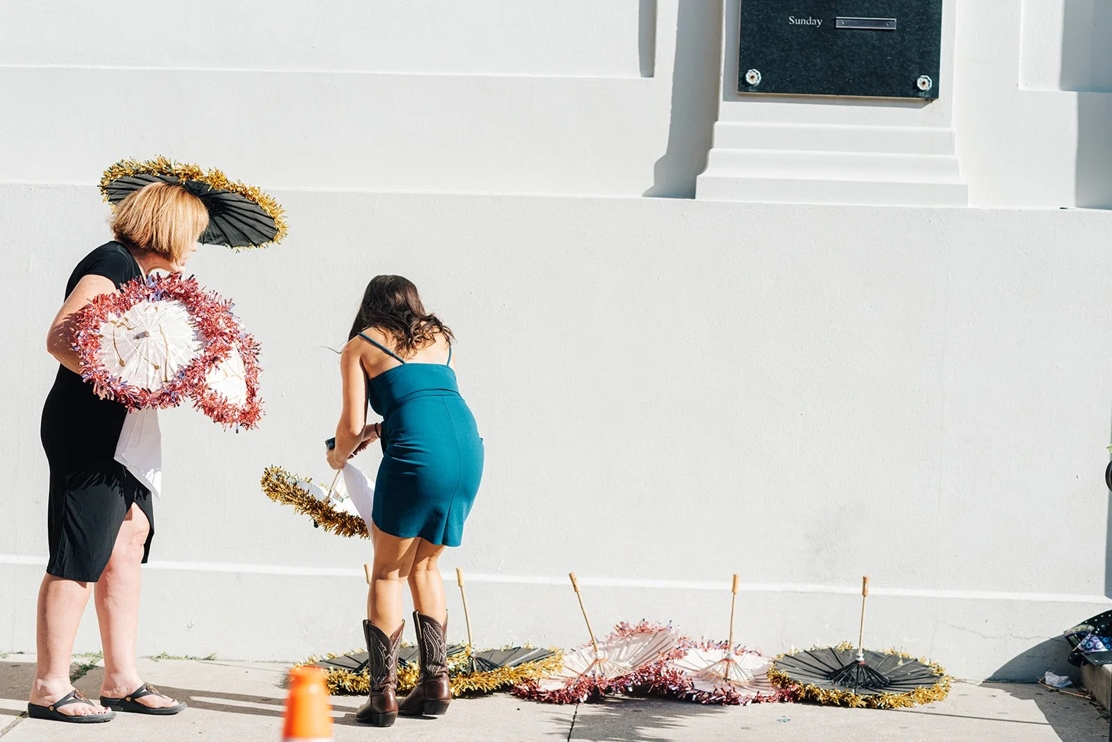 Two women, one older with short blonde hair and a black dress, and one younger with long dark hair and a blue dress, standing next to colorful parasols decorated with tinsel, on a sidewalk against a white wall