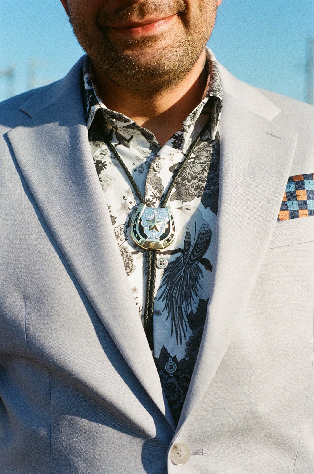 Close-up of a man wearing a light-colored blazer, a floral shirt, and a sheriff badge necklace, with a blue sky in the background.