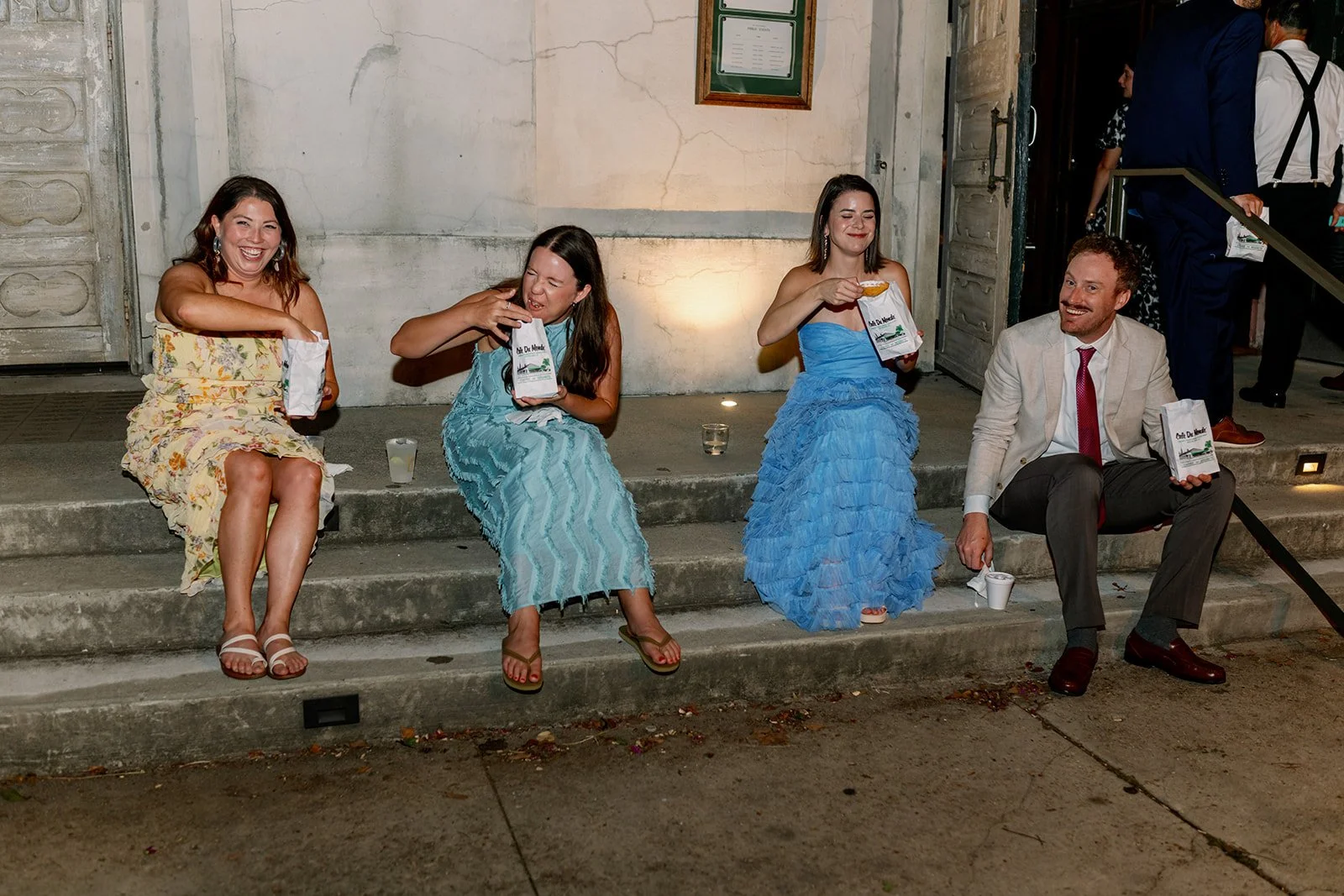 Four people sitting on steps outside at night, three women in blue dresses holding gift bags, and a man in a white blazer and red tie smiling, all enjoying a casual moment.