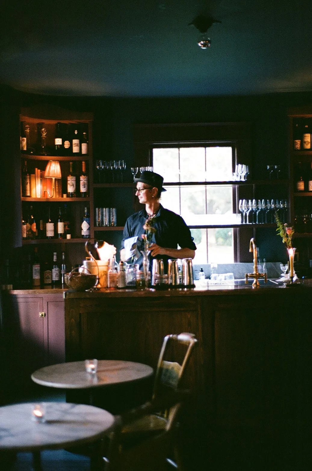 A woman bartender in black attire and a hat standing behind a bar counter with bottles, glasses, and barware, illuminated by natural light from a window behind her, in a dark, cozy bar setting.