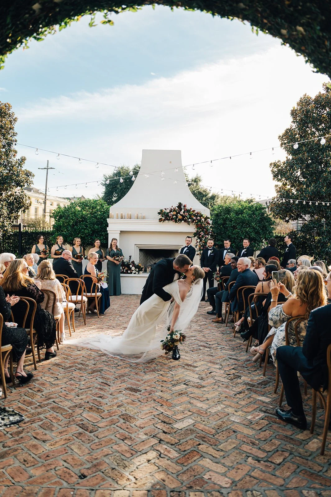 Couple kissing at their outdoor wedding ceremony, surrounded by seated guests, with bridesmaids and groomsmen standing behind a white outdoor fireplace decorated with flowers, under a sky with string lights.