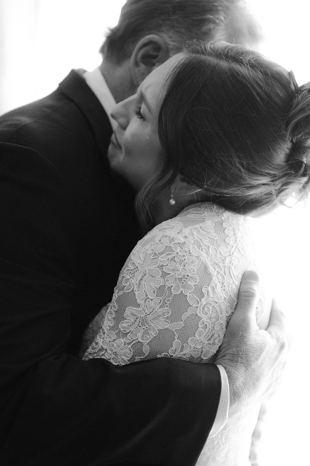 A black and white photograph of a couple sharing a kiss, with the man dressed in a suit and the woman in a lace dress. The woman has her eyes closed and is wearing a pearl earring. The man's hand is gently resting on the woman's shoulder.