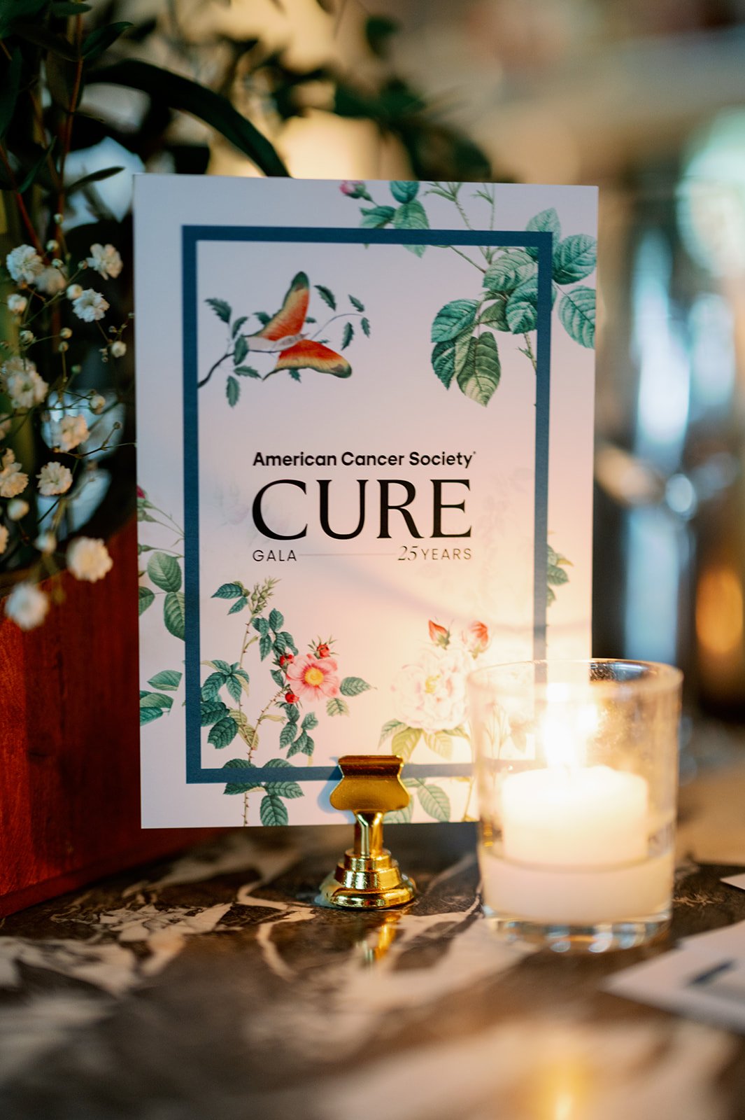 A sign for the American Cancer Society CURE Gala, celebrating 25 years, decorated with floral and bird illustrations, placed on a table with a candle in a glass holder.