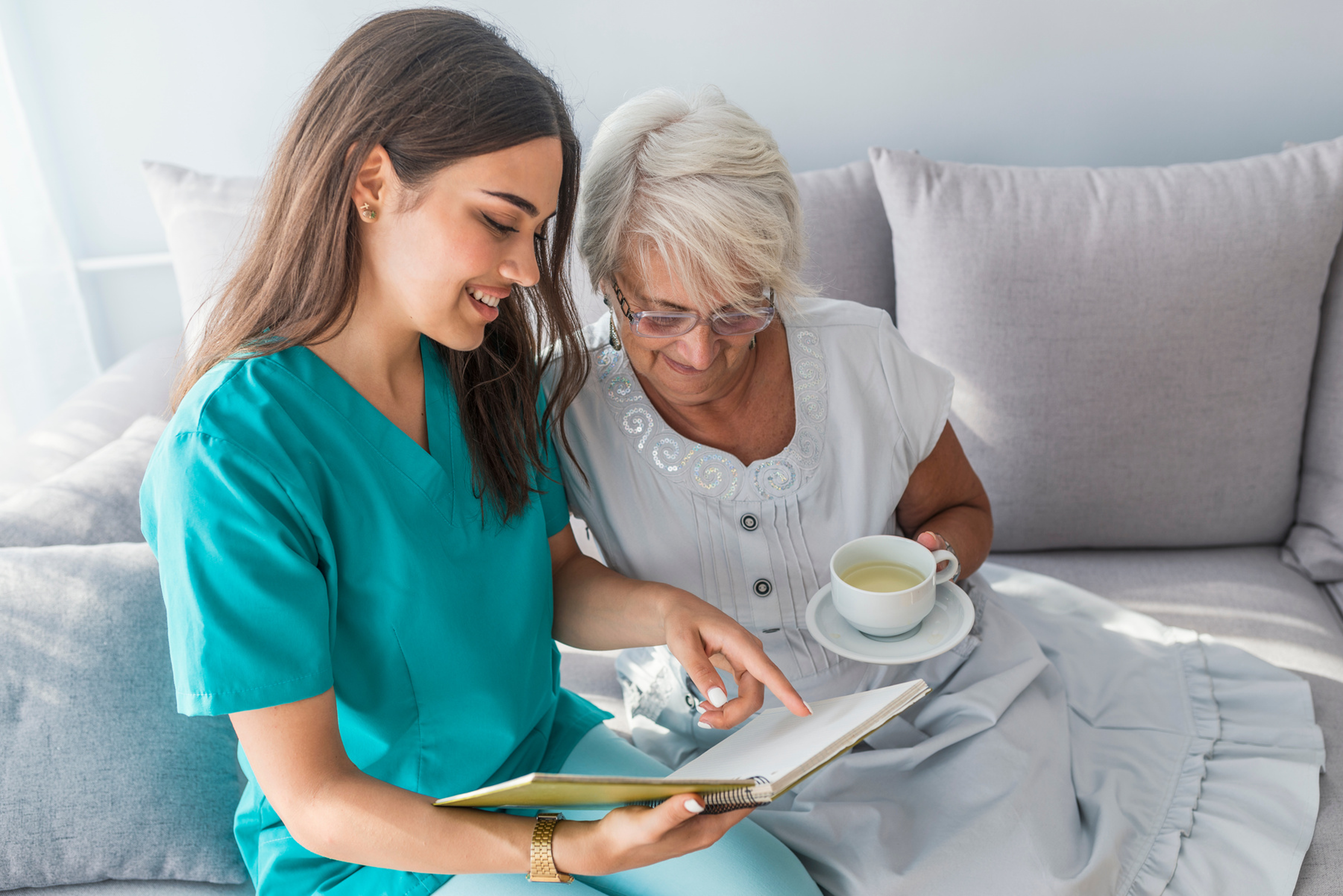 A young female nurse in teal scrubs showing an open notebook to an elderly woman with white hair and glasses, who is holding a cup of tea and sitting on a light gray couch in a well-lit room.