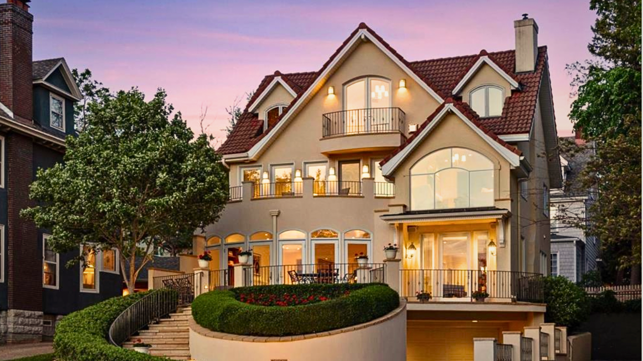 Large, multi-story house with a red-tiled roof, large windows, and balconies, illuminated at sunset, with a landscaped front yard and trees. Lake Minnetonka Luxry home. Nick Sundahl Realtor