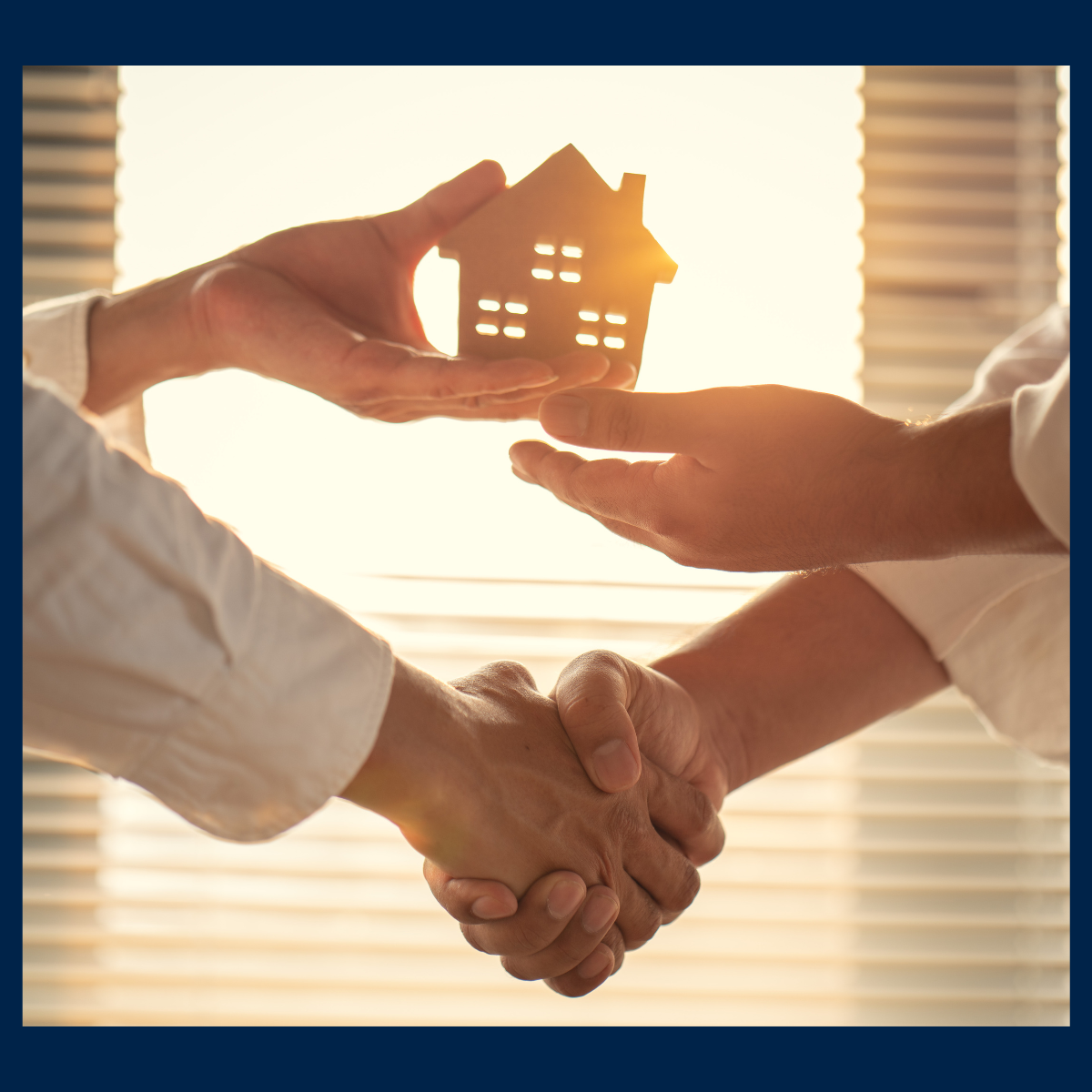 Two people shaking hands with a small house model being held above them, backlit by sunlight through window blinds.