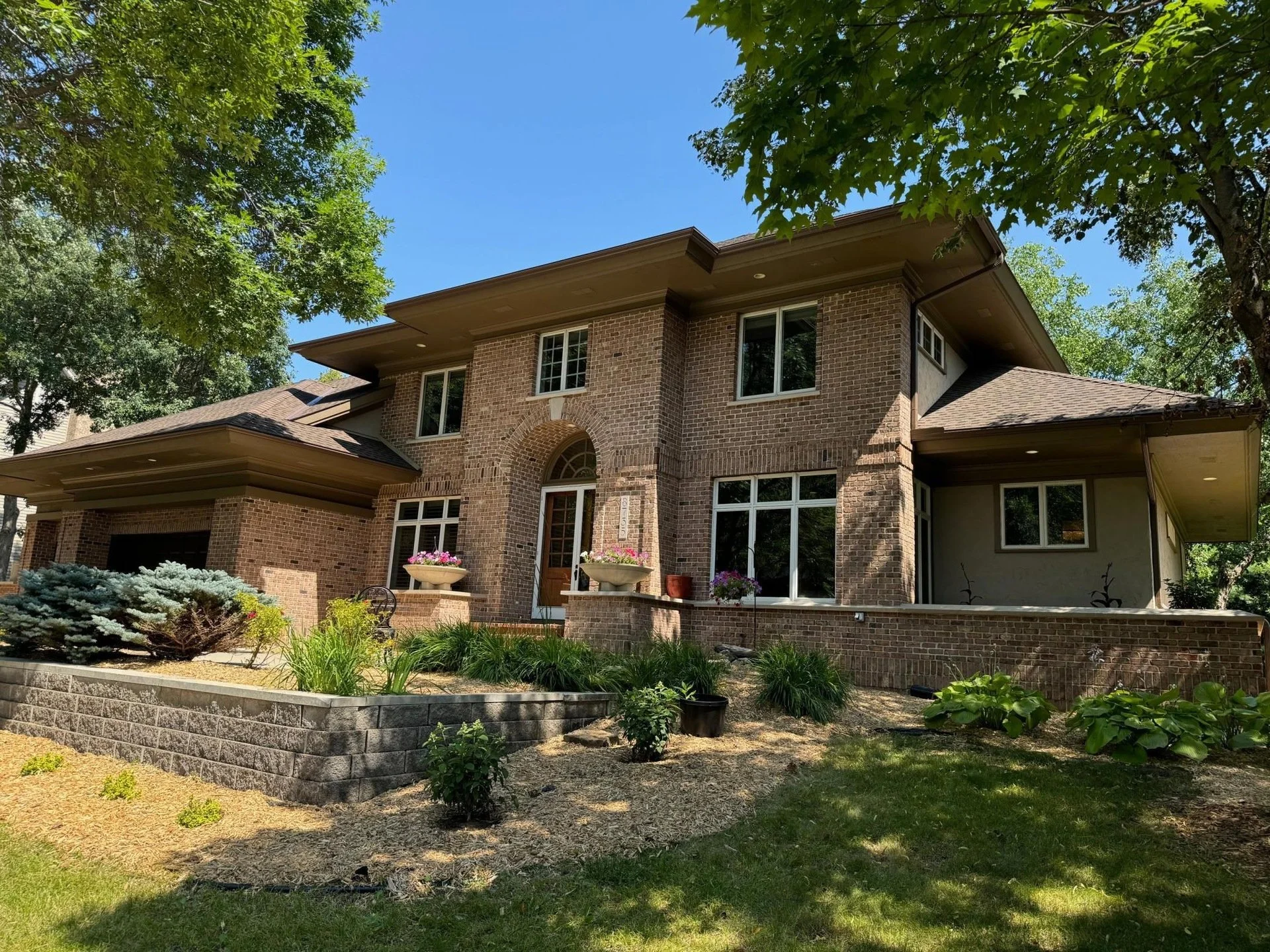 Two-story brick house with a landscaped front yard, trees, and plants. Clear blue sky in the background. Eden Prairie Luxury home. Sold by Nick Sundahl, Lakes Sotheby's International Realty
