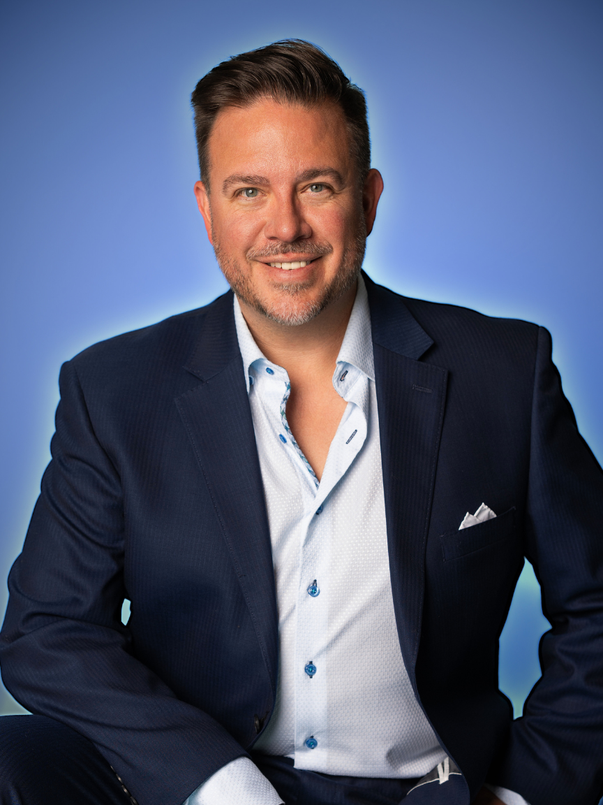 A man wearing a dark blue suit and a light blue dress shirt, smiling against a blue background. Nick Sundahl, Realtor - Lakes | Sotheby's International Realty